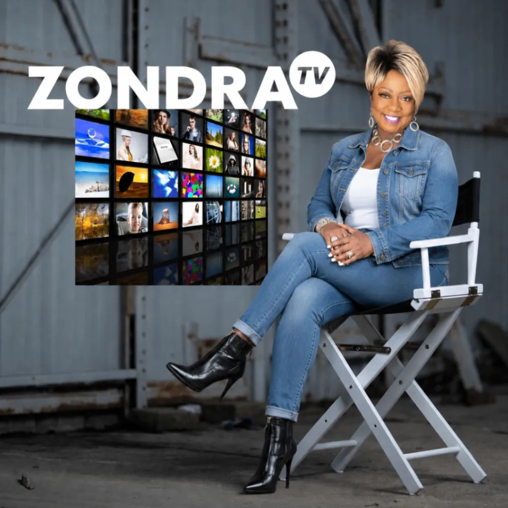 Zondra Evans seated in a director's chair in front of a wall of television screens